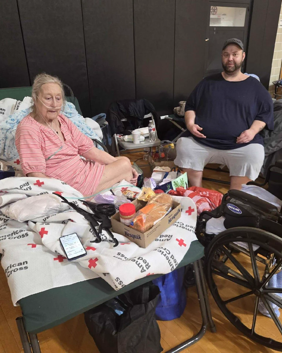 An individual sits on a cot with American Red Cross blankets, surrounded by belongings and supplies. Next to the cot, another person is seated on a chair. A wheelchair is nearby. They are in a shelter.