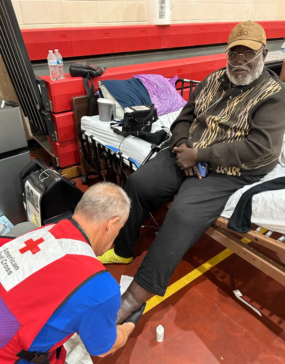 A person wearing a Red Cross vest is attending to another individual sitting on a cot. The seated person is wearing a patterned vest and cap. A bottle of lotion and some supplies are nearby. The setting appears to be a gymnasium, with cots and bottled water visible.