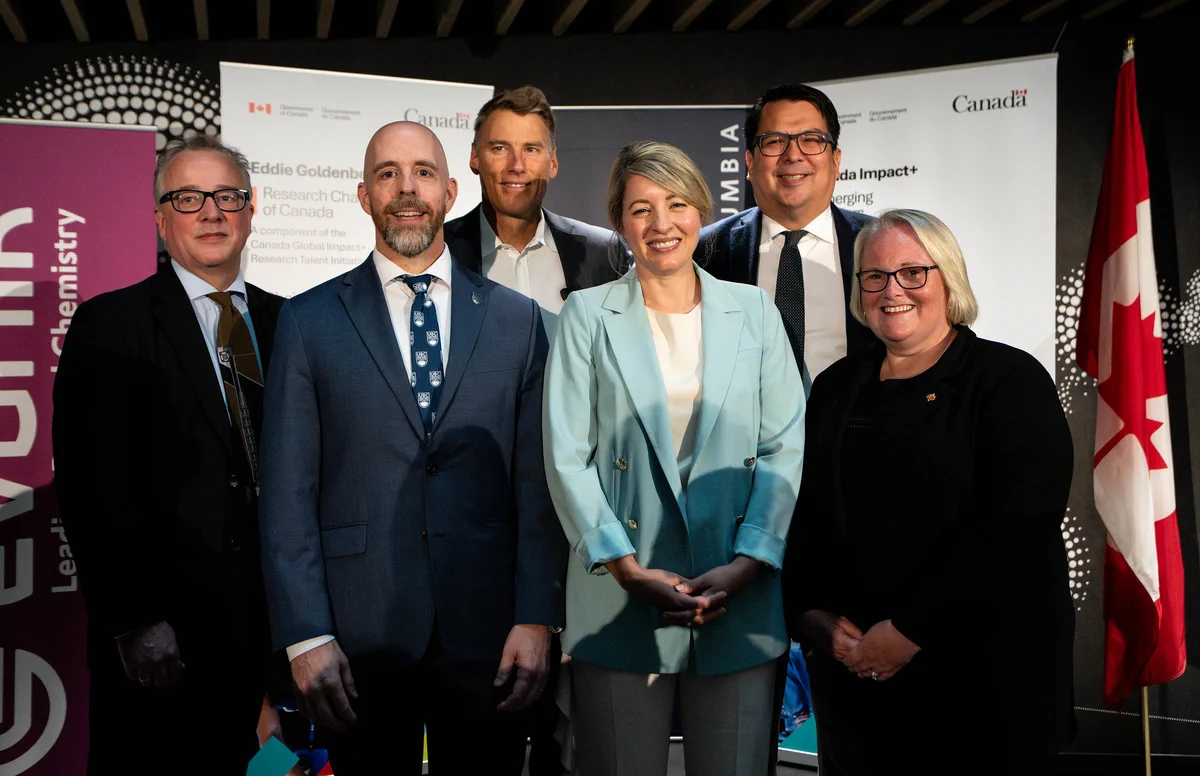 Minister Joly and five researchers posing in front of Canadian government banners and a flag, dressed in business attire at an official announcement event.