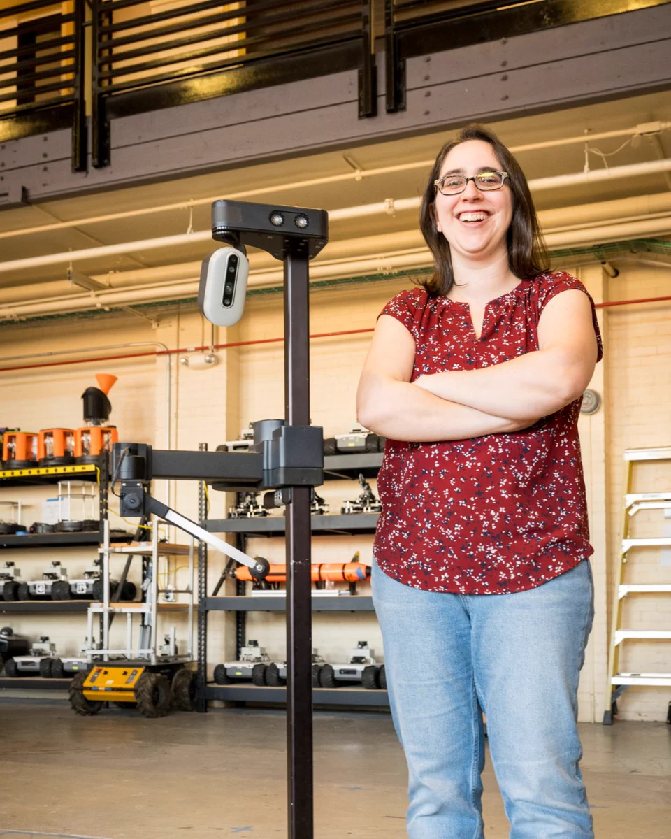 Naomi Fitter, P stands smiling beside a tall robotic structure in a lab.