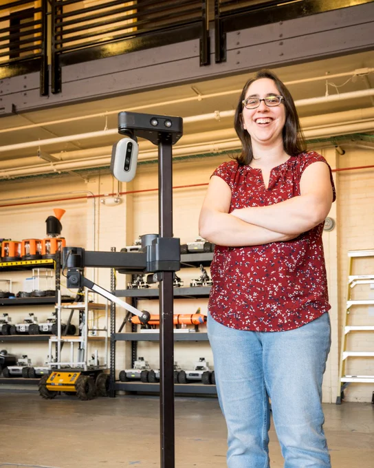 Naomi Fitter, P stands smiling beside a tall robotic structure in a lab.