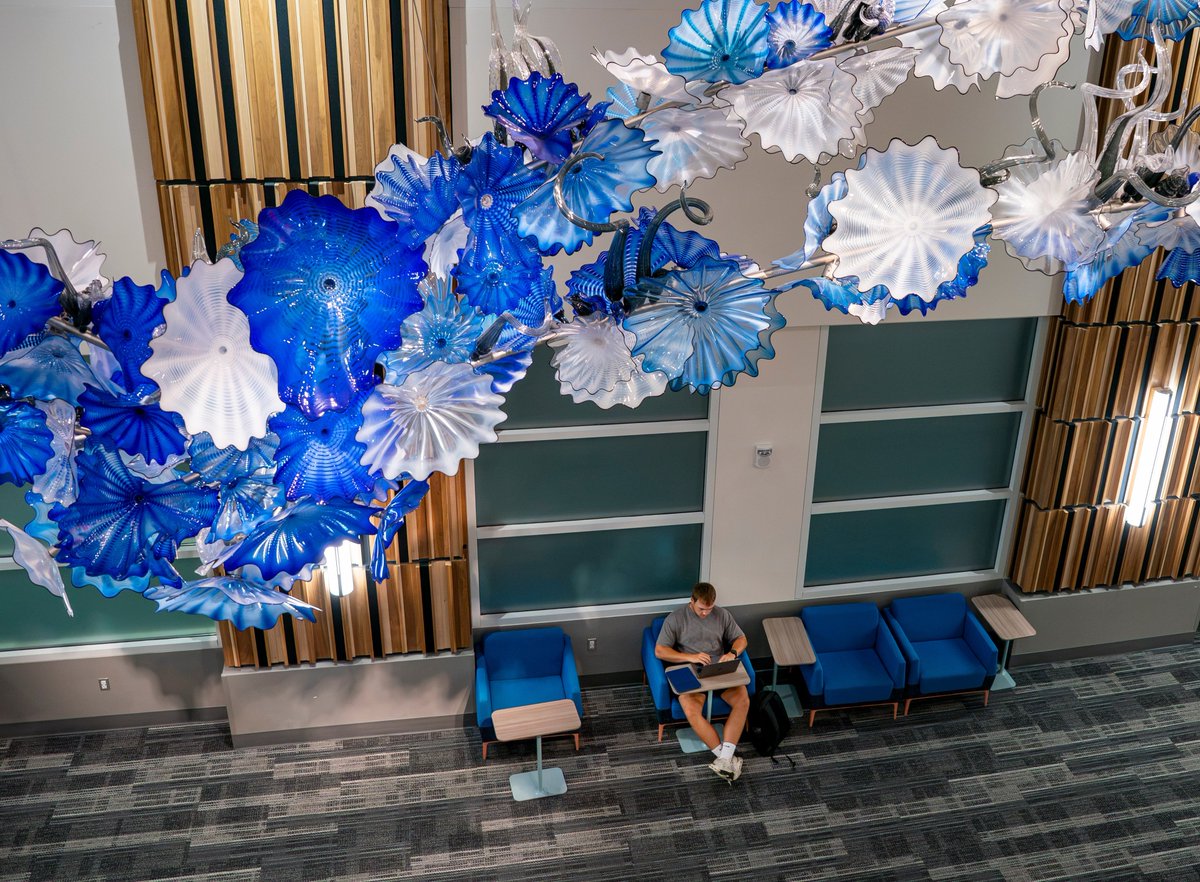 A man its in a chair below an ornate blue and white class Chihuly sculpture hanging from the ceiling.