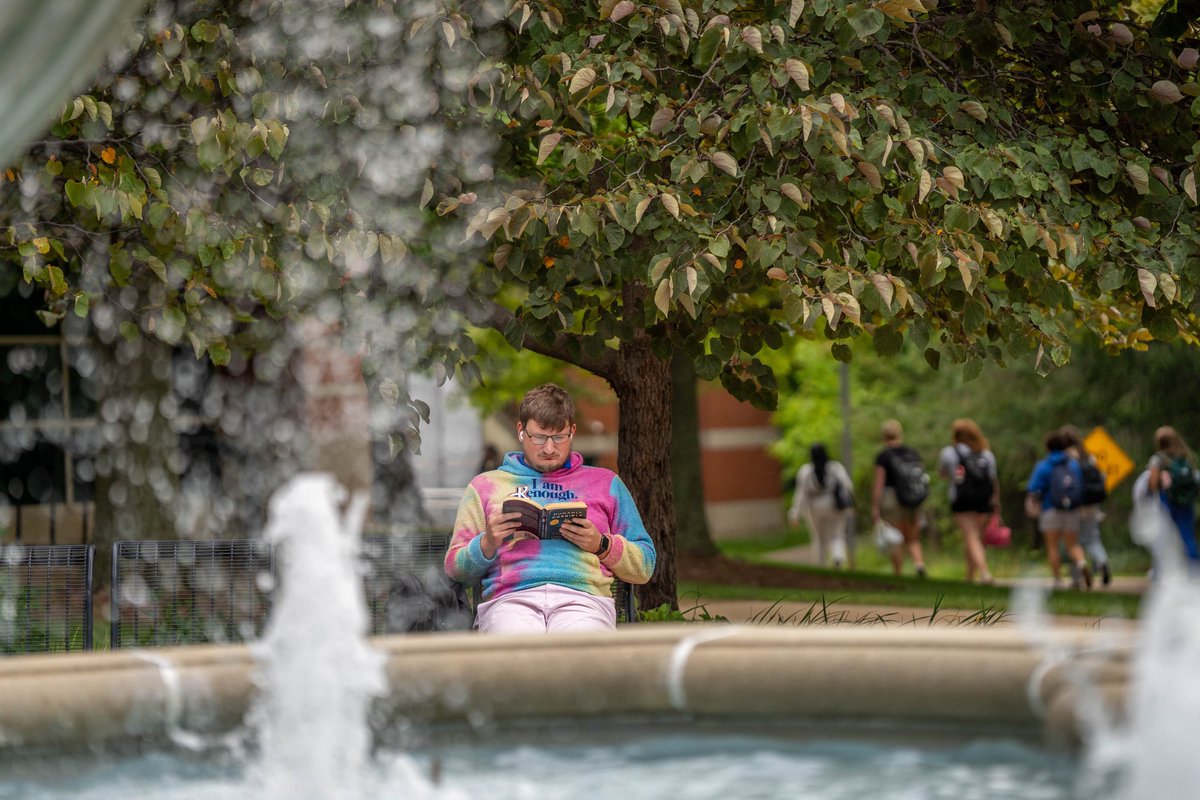 A man wearing a colorful hoodie sits near a fountain on campus and reads a book.