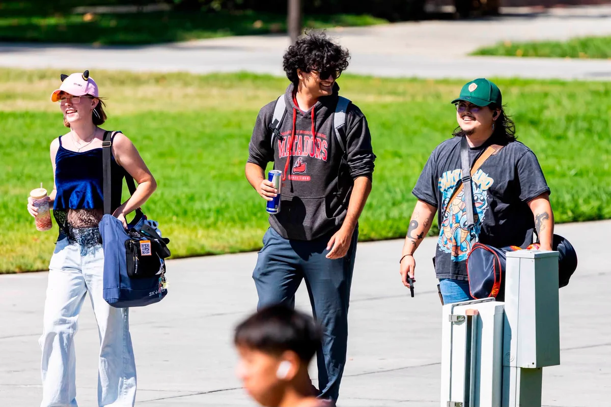 Three students walk together across the CSUN campus on a sunny day, smiling and carrying drinks and bags.
