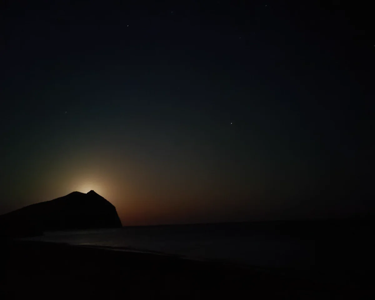 Moon rising behind the monolith at Anafi, casting a warm glow across a dark calm sea beneath a deep, star-speckled night sky.