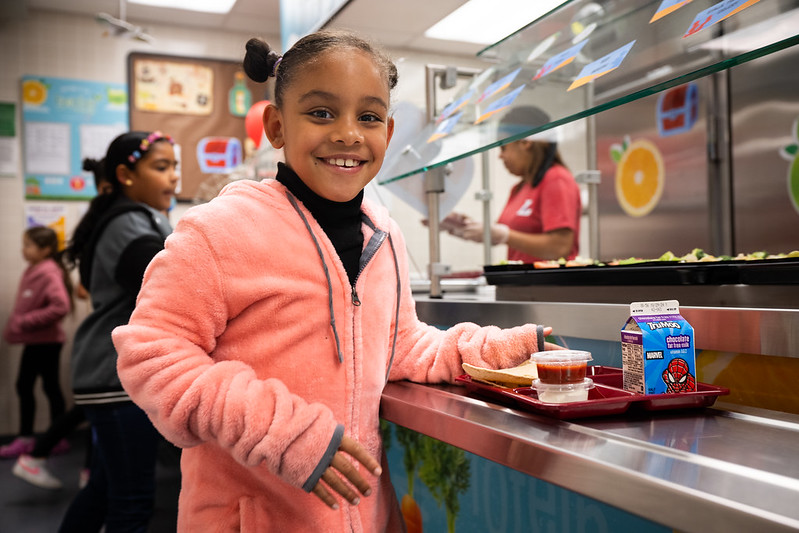A young girl standing in school lunch line smiling.