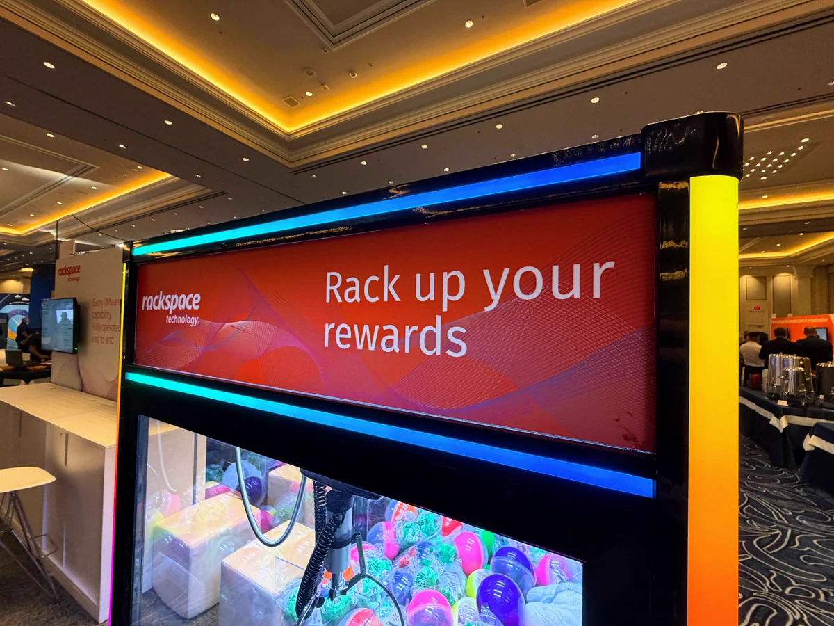 A Rackspace Technology branded claw machine at an event, with "Rack up your rewards" displayed on the top. The machine contains colorful balls. Event tables and lights are visible in the background.