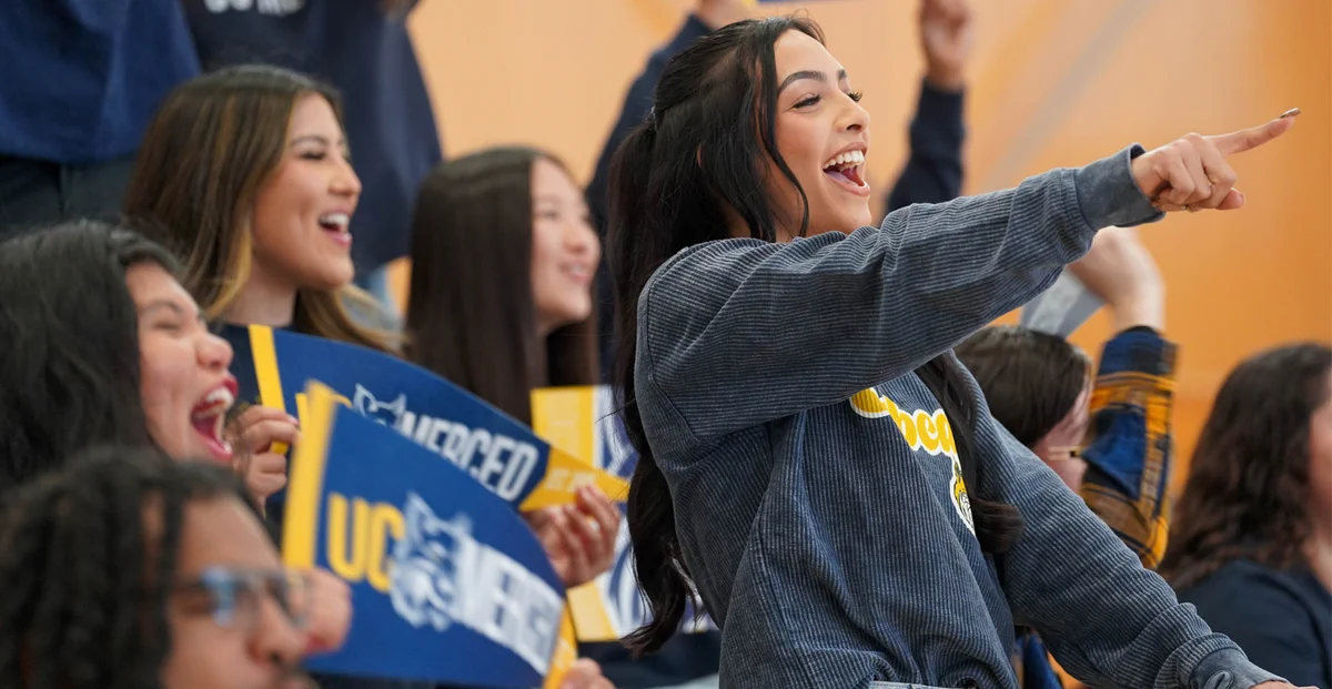 Excited young woman enthusiastically cheering and pointing during a lively sports event with fellow fans holding blue and yellow signs.