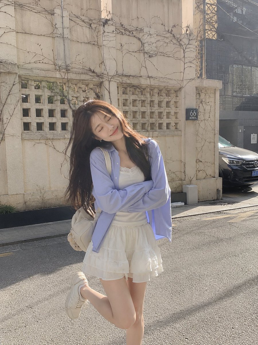 Aqsahere_'s tweet photo. Chatgpt image 2 

Prompt:
A photorealistic full-body outdoor portrait of a young woman standing on a quiet urban street in soft winter sunlight. She has long, straight dark-brown hair flowing naturally over her shoulders and a gentle, cheerful expression with her eyes softly closed. Her head is tilted slightly to one side, creating a playful candid pose.
She wears an oversized pastel lavender-blue button-up shirt layered over a delicate white lace-trimmed top, paired with a short flowing white tiered skirt. A small cream-colored backpack hangs over one shoulder. She stands with her arms comfortably crossed while lifting one leg slightly behind her, wearing white sneakers.

The background features an old textured cream-colored wall with geometric square details, bare climbing vines, a small dark sign, a parked car, and an urban street setting. Soft afternoon sunlight creates a subtle warm glow around her hair, gentle shadows, and a faint lens flare.
Natural smartphone photography, candid travel-fashion aesthetic, realistic skin texture, detailed hair strands and fabric, soft pastel color grading, shallow depth of field, subtle vintage film grain, natural proportions, dreamy peaceful atmosphere, highly photorealistic, vertical 4:5 composition.