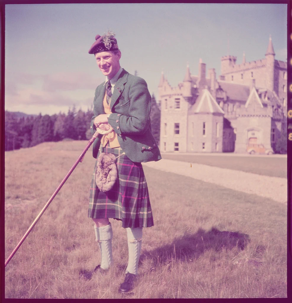 A man in a green coat, kilt and hat stands in front of a castle. September, 1950. Toni Frissell.