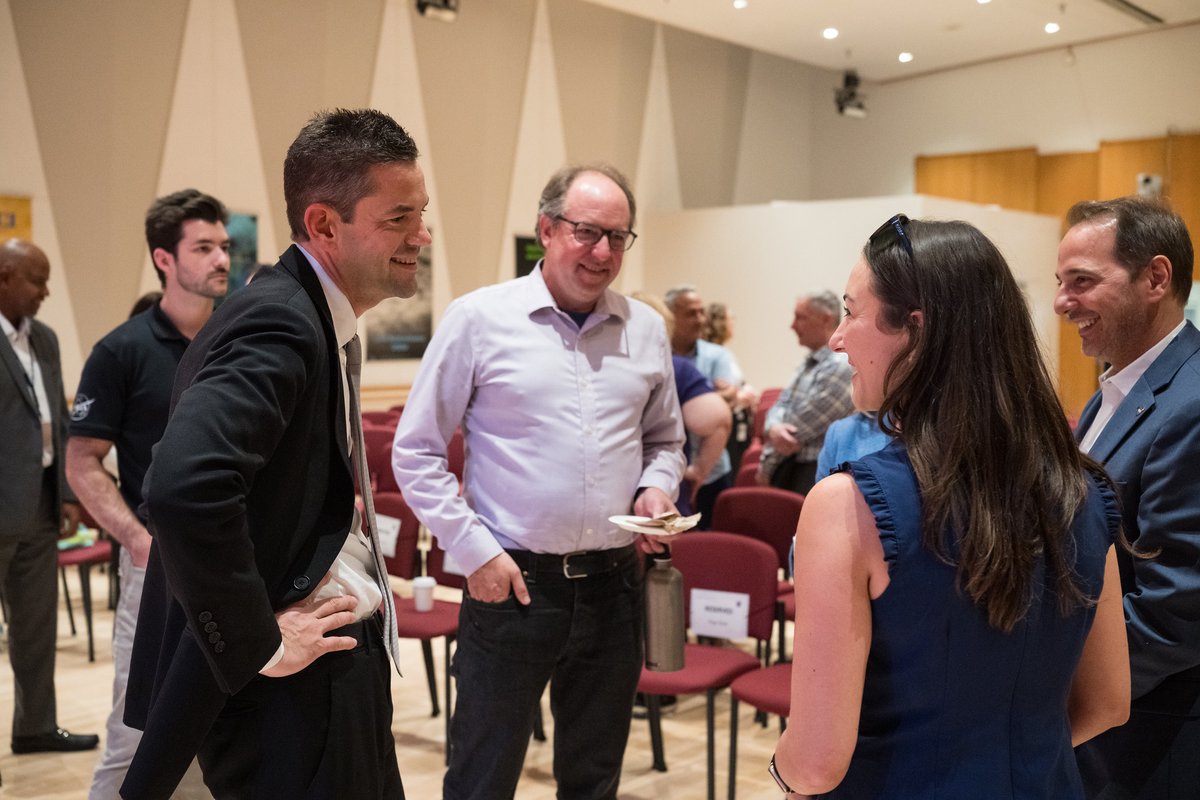 Jared Isaacman listens to an employee during an informal meet and greet. They are standing facing each other and smiling. Tom Neumann stands next to Jared, also smiling. They are in a well-lit auditorium.