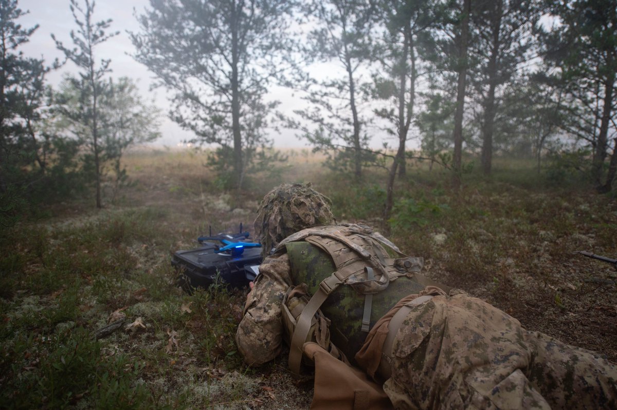 A soldier lying prone on forest floor operating a drone in a misty woodland setting.