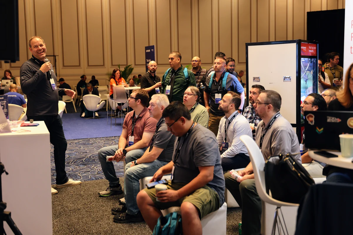 A person stands and speaks to a seated group at a conference. Attendees show interest in the presentation. The backdrop includes a wall with vertical panels, and the atmosphere appears professional and engaged. People have notebooks and some are holding refreshments.