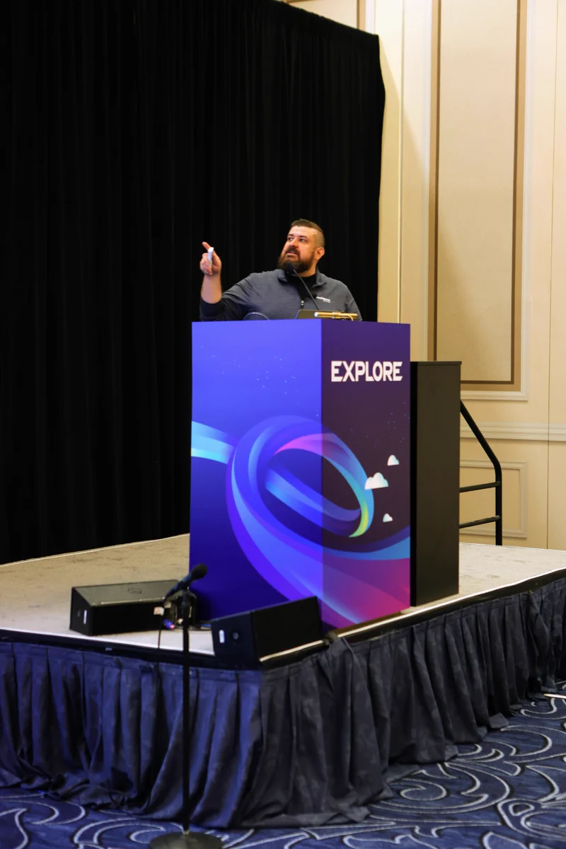 A person stands at a podium labeled "Explore" on a stage with a dark curtain backdrop. They are gesturing upwards with one hand. The floor is carpeted with a wave-like pattern.