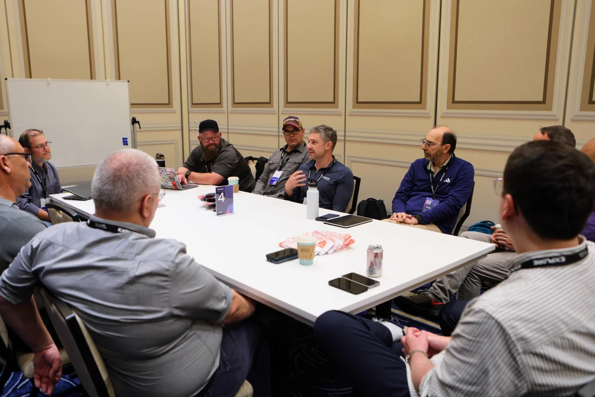 A group of people are seated around a table in a conference room, engaged in discussion. Various items like drinks, bags, and notebooks are on the table. The setting appears informal, yet focused. A whiteboard is visible in the background.