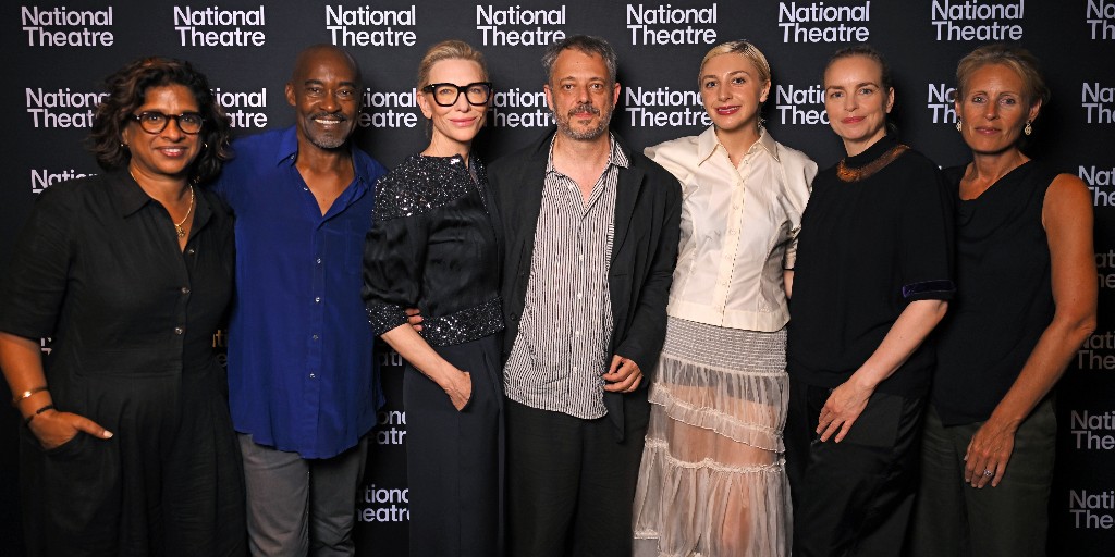 The cast and creatives of Electra/Persona smile and stand together in front of a photo wall tiled with the National Theatre logo. They are dressed formally and festively in an array of buttoned shirts to long skirts.