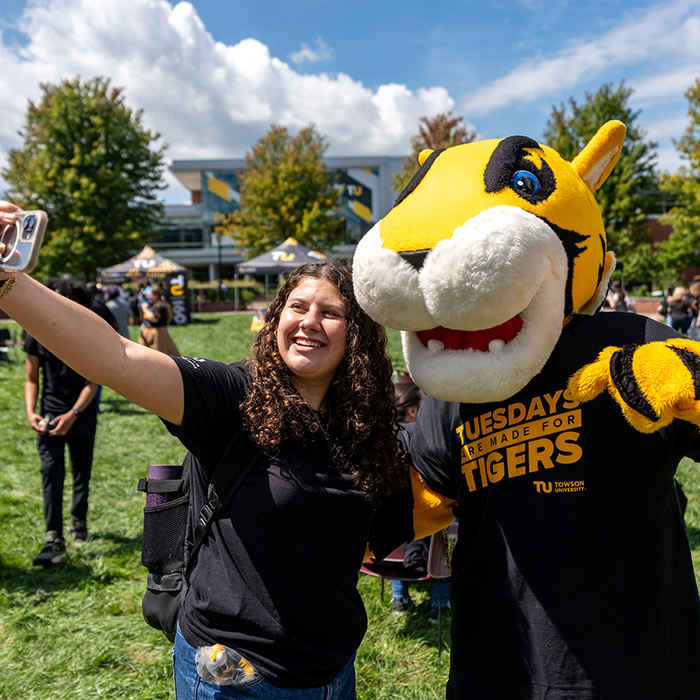 A person is taking a selfie outdoors with the Towson University tiger mascot. Behind them, there are trees, people, and a building in the background. The mascot is wearing a T-shirt with the words "Tuesdays are made for Tigers" and the Towson University logo.