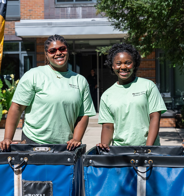 Two people in light green T-shirts are standing outdoors with large blue carts, smiling at the camera. They appear to be engaging in an event or activity outside a building.