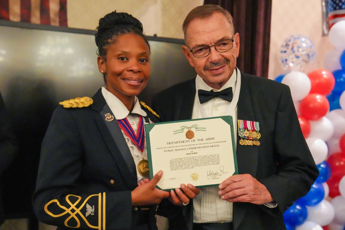 Two people in military uniform stand inside during a formal even with balloons in the background. One person is handing the other a certificate for a Public Service Commendation Medal.
