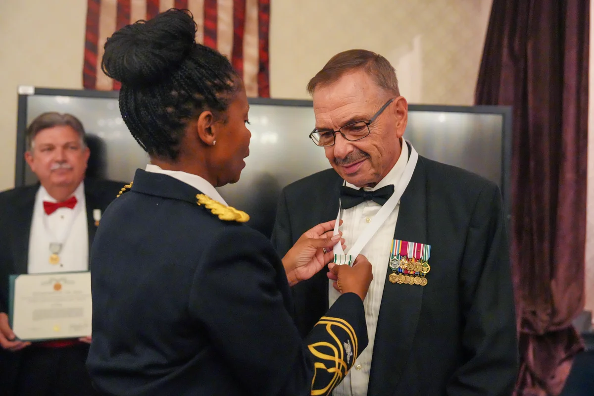 A person in a military uniform is awarding a medal to another individual wearing a formal suit with several other medals. An American flag is visible in the background, and another person is holding a certificate behind them.