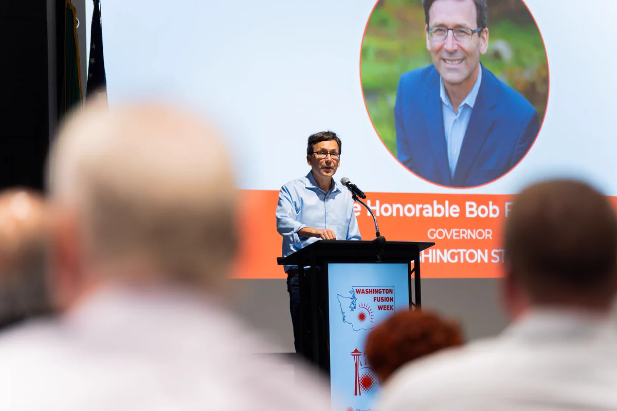 A person stands at a podium speaking, with a large screen behind displaying The Honorable Bob Ferguson, Governor Washington State and a photo. The podium features a Washington Fusion Week graphic.