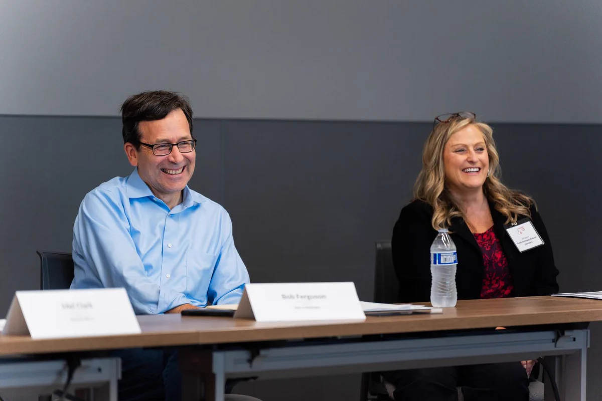 A group of individuals is seated at a conference table. Two people are prominently visible, smiling and engaged. Name tags, a water bottle and meeting materials are present on the table.