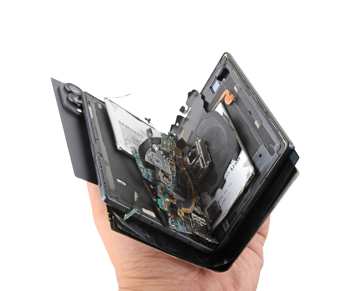 A severely damaged foldable smartphone is held open in one hand, exposing its internal components. The batteries, circuit boards, wireless charging coil, cameras, and flex cables are visible between the separated and distorted sections of the device.
