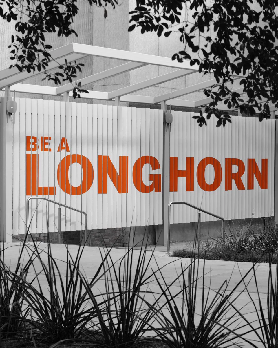 A white slatted fence displays bold orange text: 'BE A LONGHORN'. The rest of the scene is grayscale, including a building facade, an overhead structure, dark tree foliage at the top, and spiky plants on concrete ground with metal handrails.