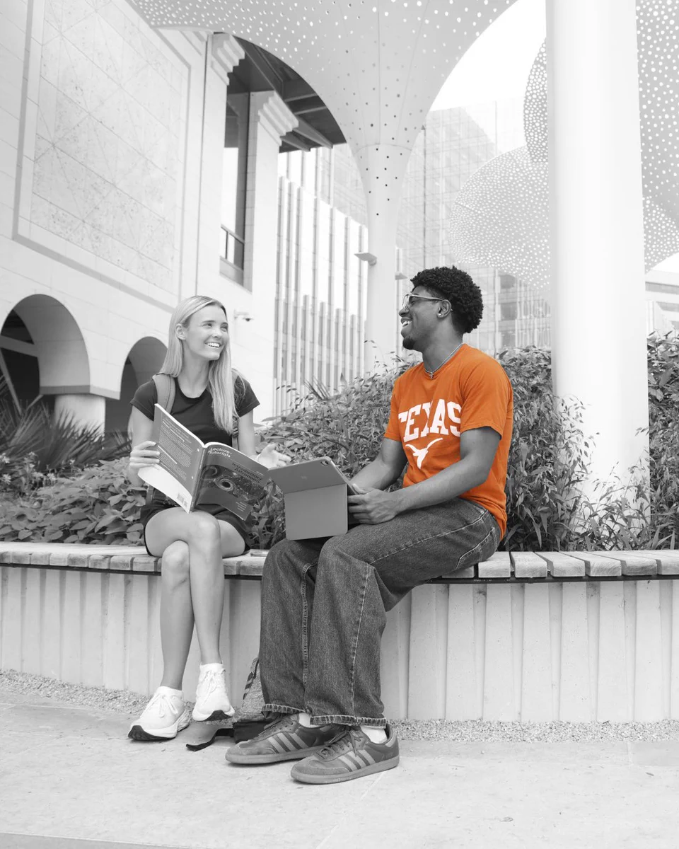 Fair-skinned woman with blonde hair and dark-skinned man with curly hair and glasses sit on a bench, smiling and talking. She holds a textbook, he holds a tablet and wears an orange \"TEXAS\" shirt. Desaturated modern outdoor background.