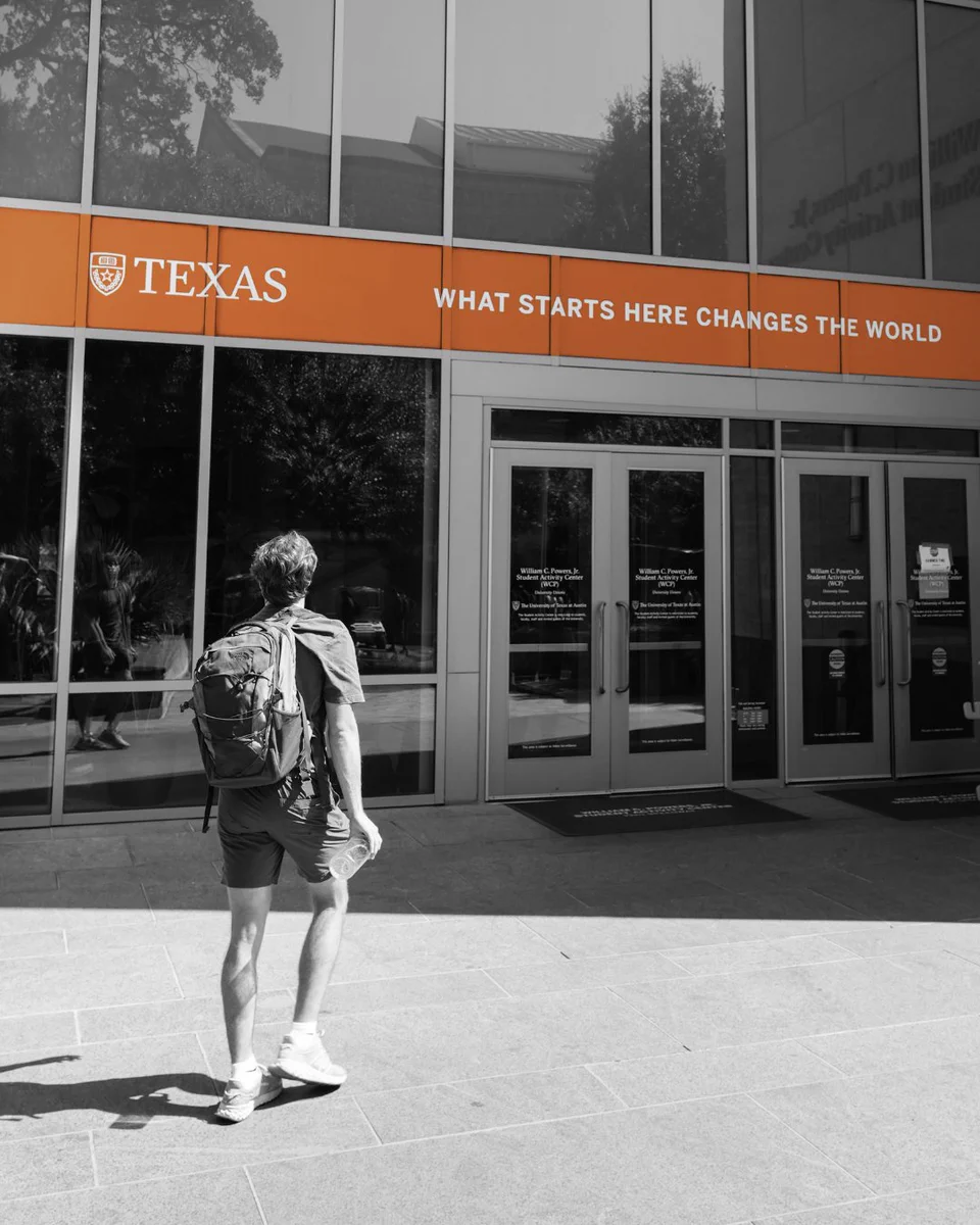 A person with a backpack walks toward a building with an orange banner that reads \"TEXAS WHAT STARTS HERE CHANGES THE WORLD\". The person and building are in black and white, while the banner is in color. Doors read \"William C. Powers, Jr. Student Activity Center (WCP)\".