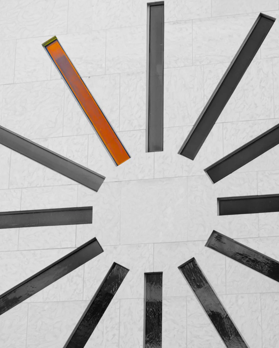 A light tiled wall with dark gray and black rectangular bars radiating outwards, forming a circle. One bar at the top-left is distinctly bright orange, standing out against the monochrome.