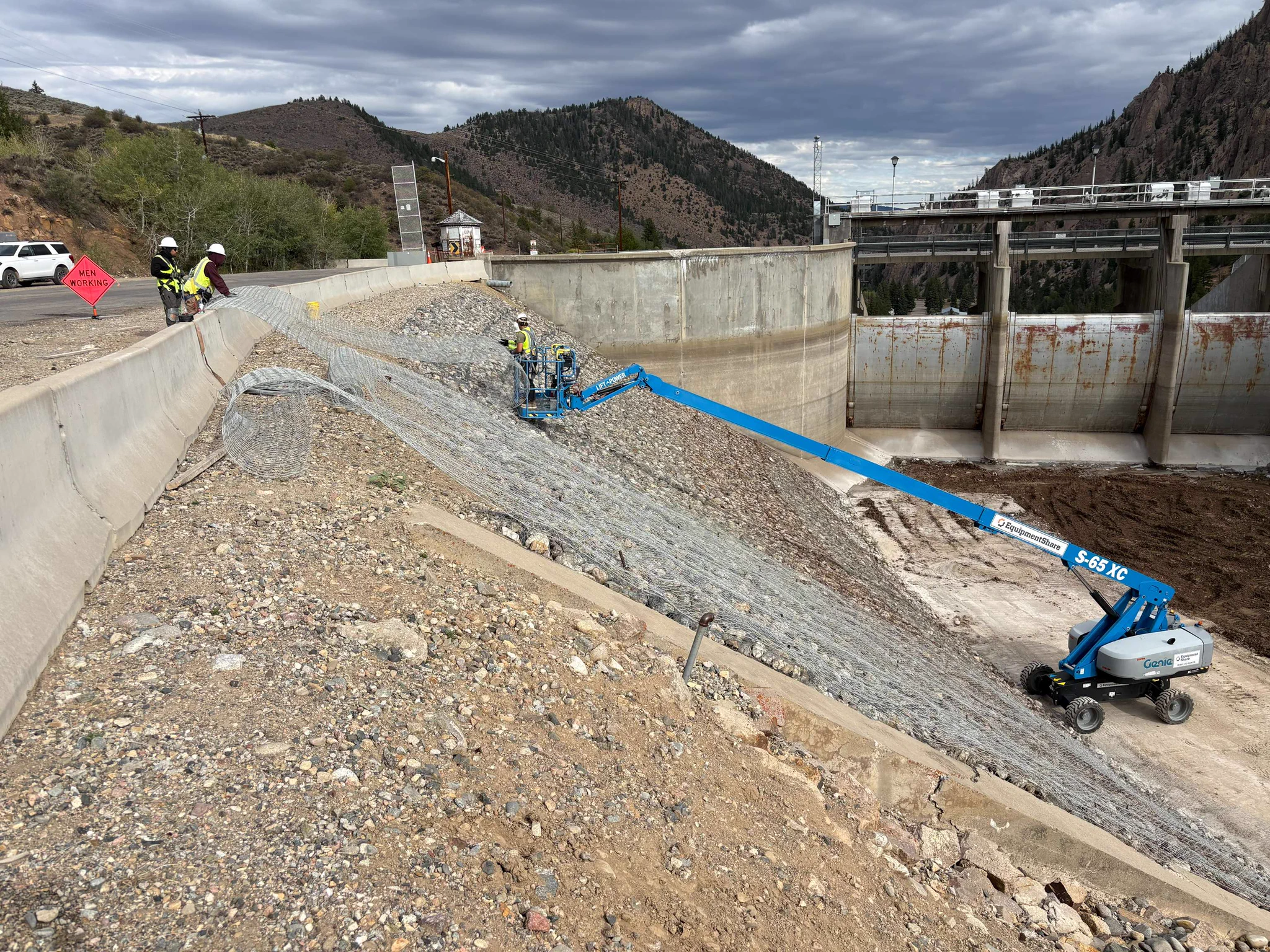 Crews install wire mesh on a large gabion adjacent to a dam spillway.