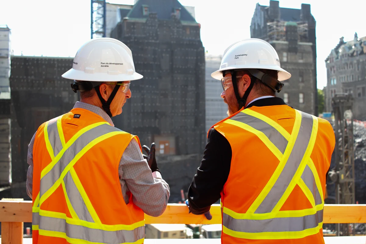 Two people wearing white hard hats, safety glasses and high-visibility orange vests stand on an elevated construction platform, looking out over a large excavation site with historic stone buildings in the background.