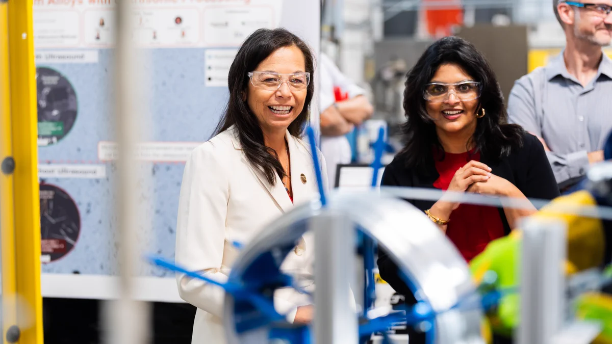 Two people wearing safety glasses are smiling and standing in a busy industrial setting with machinery and informational posters in the background.