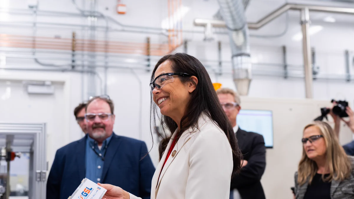 The scene shows a group of people wearing safety goggles, standing inside a lab or industrial setting with machinery and visible pipes. One person is holding a battery and smiling, while others observe.