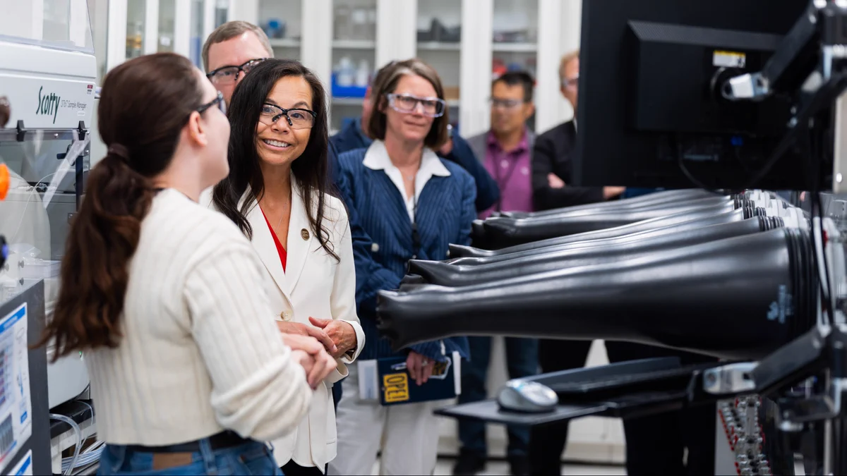 A group of people stands in a laboratory, observing a demonstration of scientific equipment with black glove ports. A person in a white blazer is engaged with others, and scientific instruments are in the background.