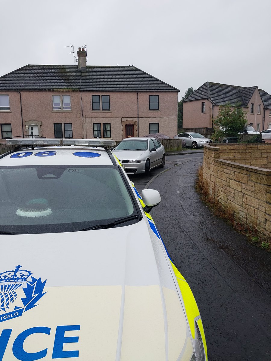 A view from a police car looking out onto a residential street with parked cars and brick houses under a grey sky.