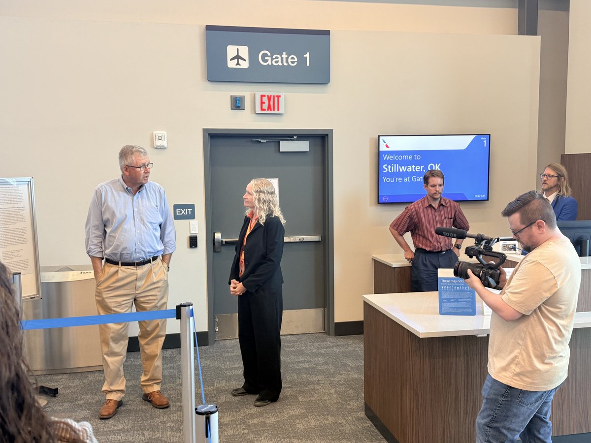 Inside an airport gate area, two people stand talking near a door while a camera operator films them. Other people and a check-in counter are visible. Signs and a screen display text.
