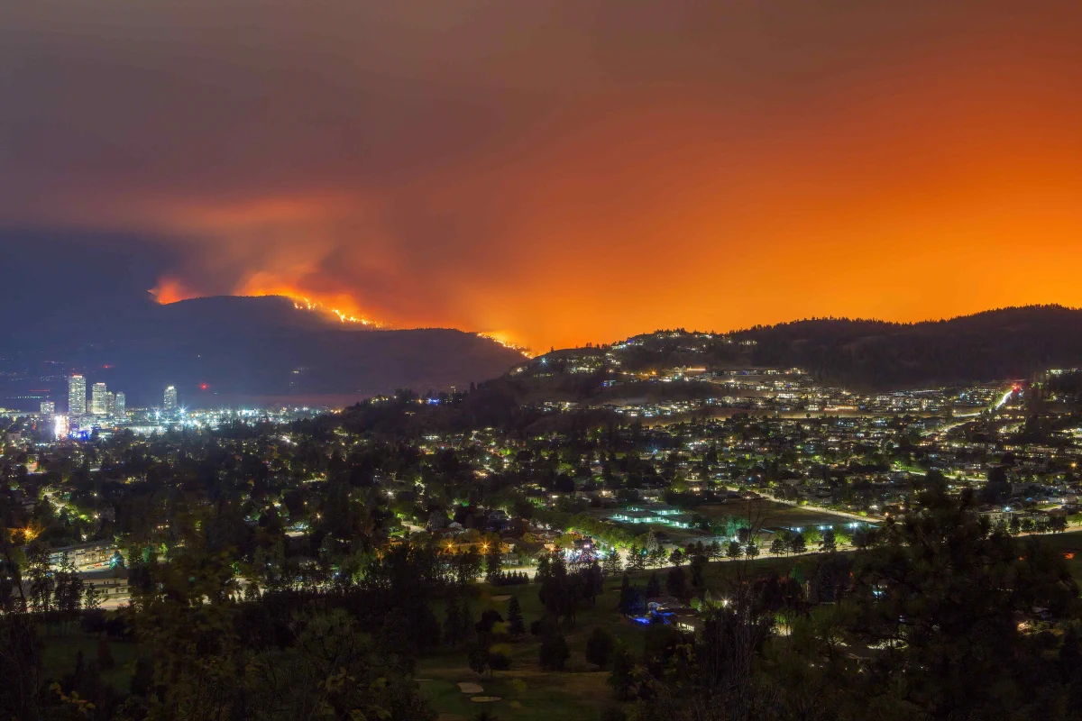 Nighttime view of a city nestled among hills as a wildfire burns along a ridgeline in the background. Bright flames stretch across the hillside, sending thick smoke into the sky, which glows orange from the fire. City lights illuminate neighborhoods, streets, and buildings below, creating a stark contrast between the urban landscape and the active wildfire.