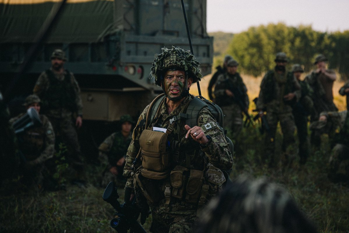 A soldier in camouflage gear and helmet is giving instructions during a field operation with troops and military vehicle in background.