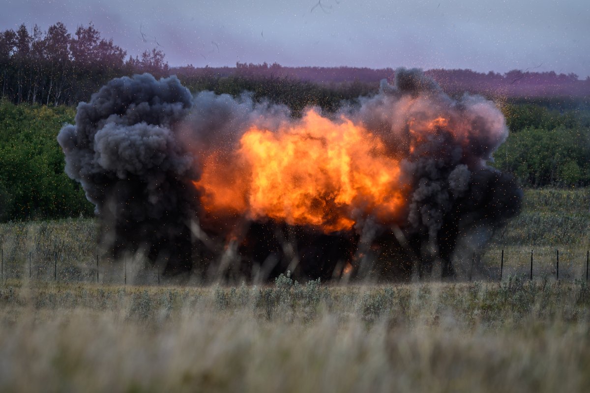 Massive explosion with bright orange flames and thick black smoke billowing over a grassy field near a forest edge.