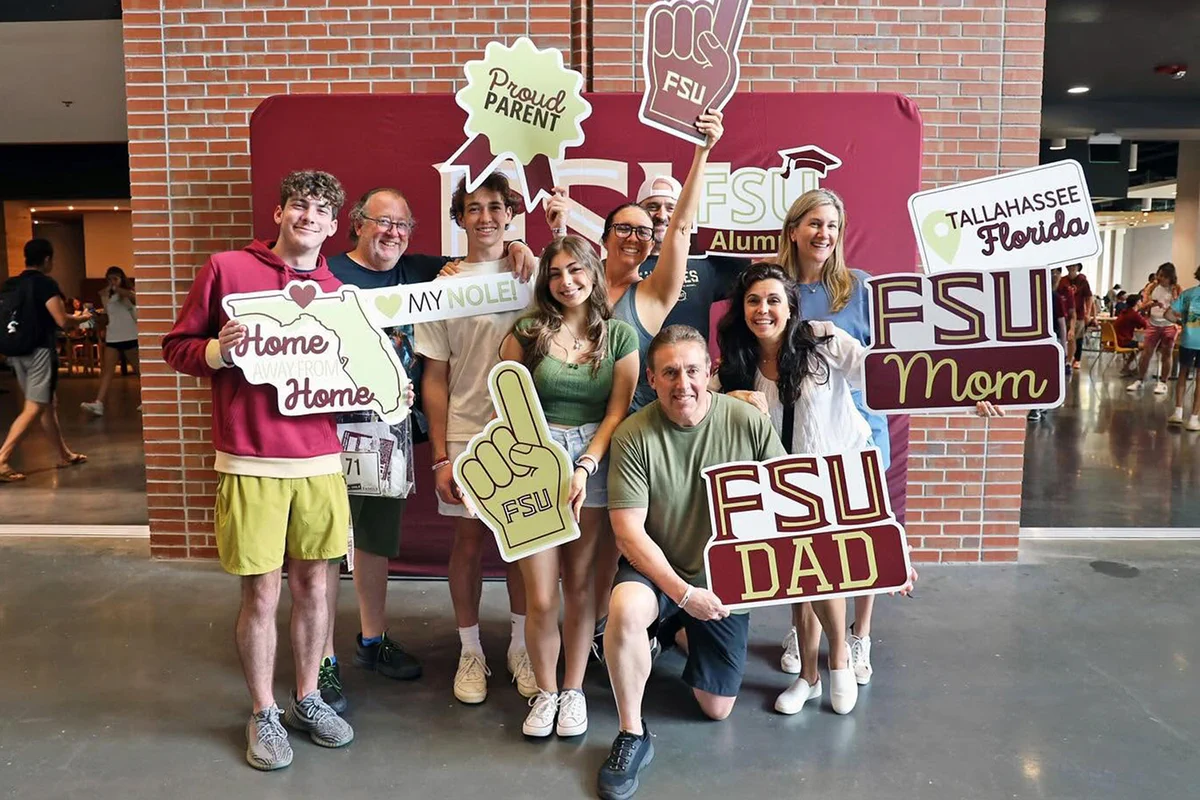 A group of eight people smile for a photo in front of a maroon Florida State University photo backdrop set against a brick wall in the FSU Student Union. They hold various handheld photo booth props, including signs reading "FSU DAD," "FSU Mom," "Proud Parent," "Home Away From Home," "My Nole," and "Tallahassee Florida," along with maroon and white foam fingers.