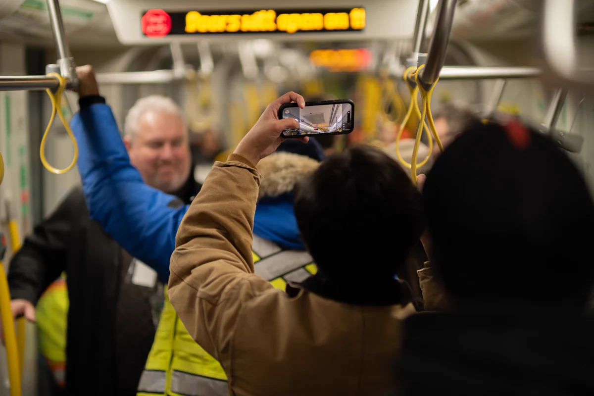 People stand on a light rail platform beside a Metrolinx train, while one person holds up a phone to take a photo. Yellow safety barriers and another train are visible in the background.