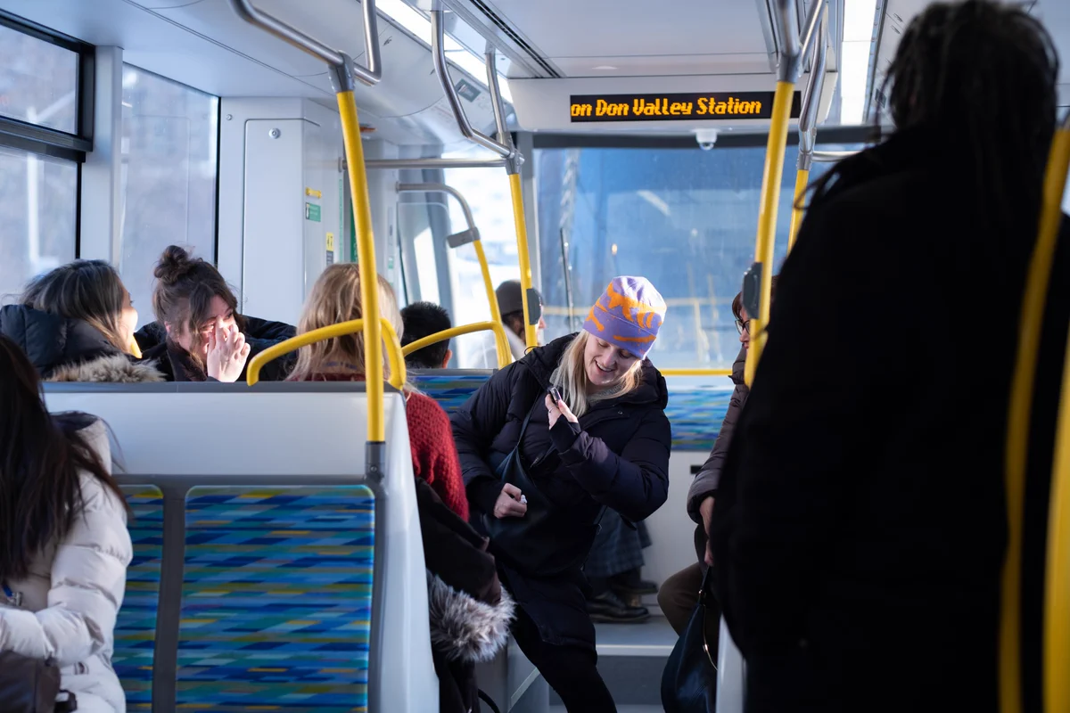 Passengers ride aboard a Line 5 train, with seated and standing riders visible inside the bright, modern vehicle as a digital display shows the approach to Don Valley Station.
