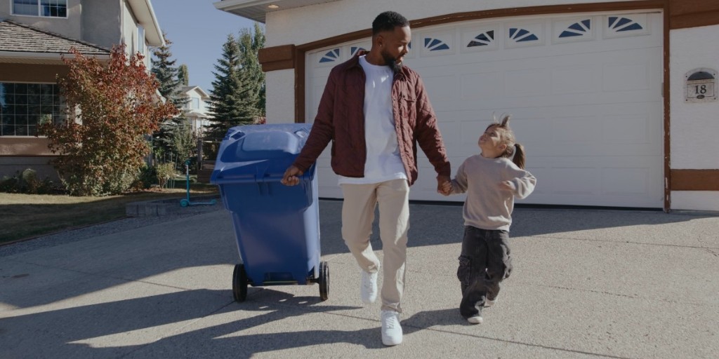 Father and young daughter holding hands while taking out a blue recycling cart on a driveway on a sunny day.