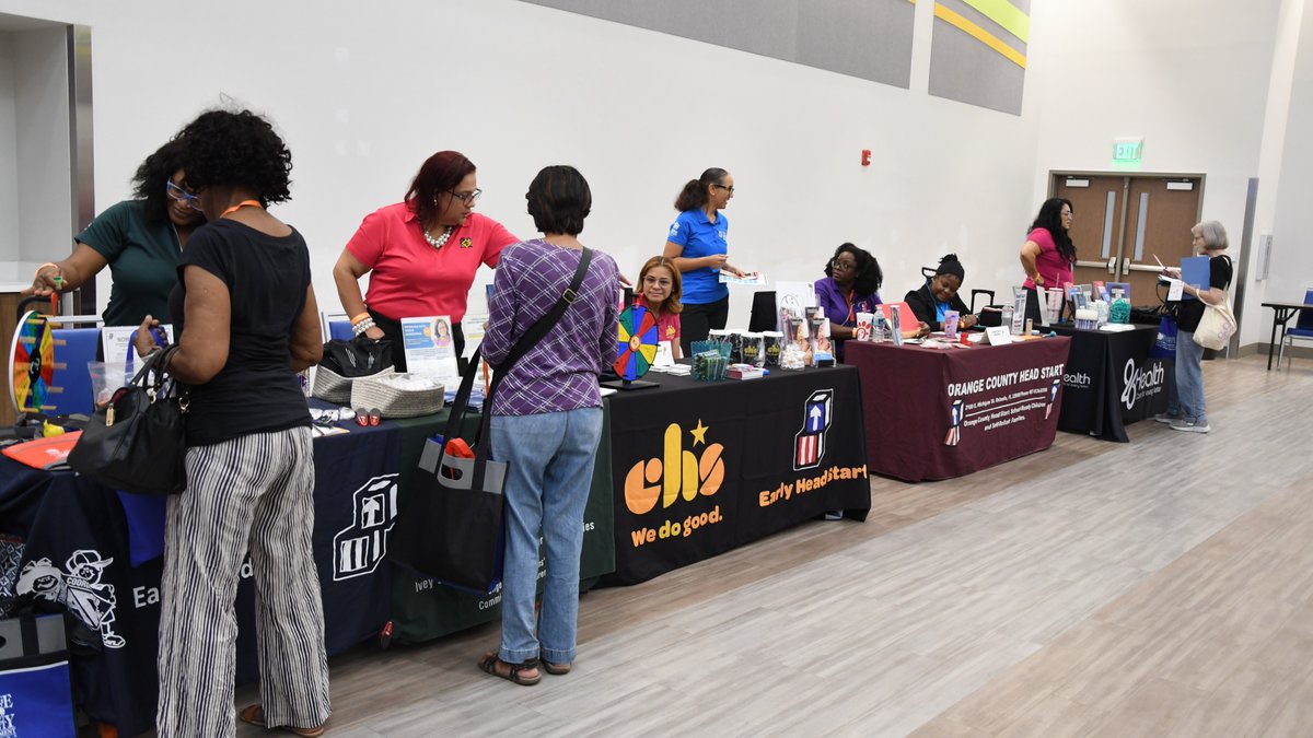 A group of people are standing and interacting at resource tables covered with various materials and promotional items. The tables have banners displaying logos and names. The event is taking place at the Orange County Multicultural Center.