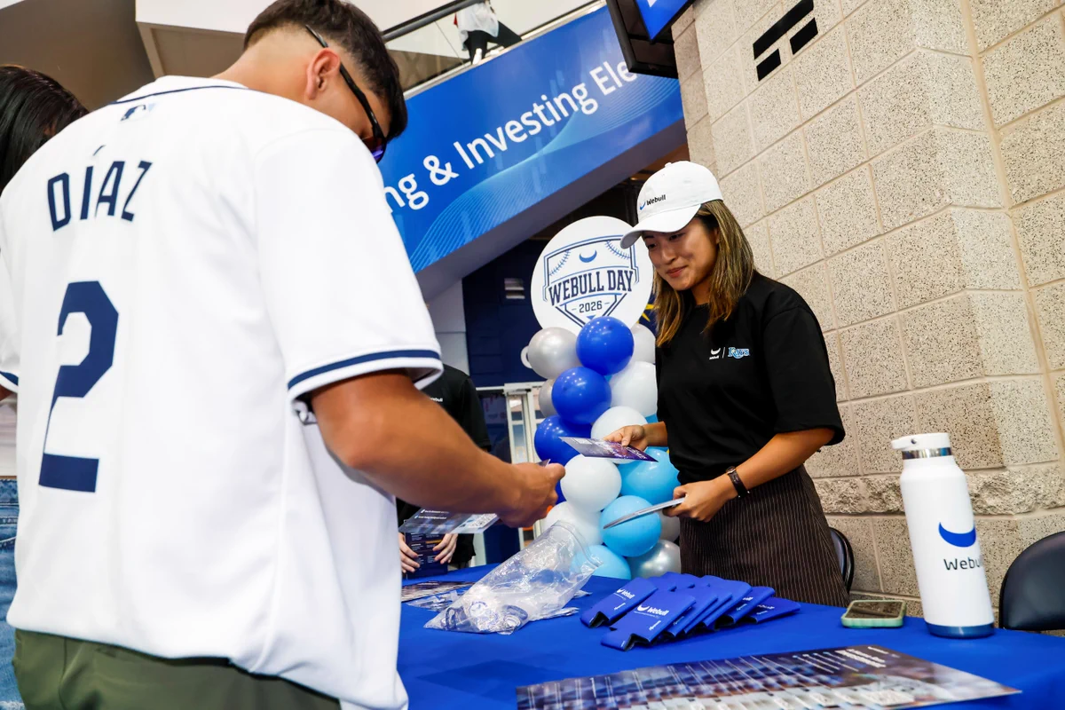 A Rays player in a "Díaz" jersey receives a card from a Webull staff member at the Webull Day 2026 merchandise table, with koozies and a blue-and-white balloon display at Tropicana Field.