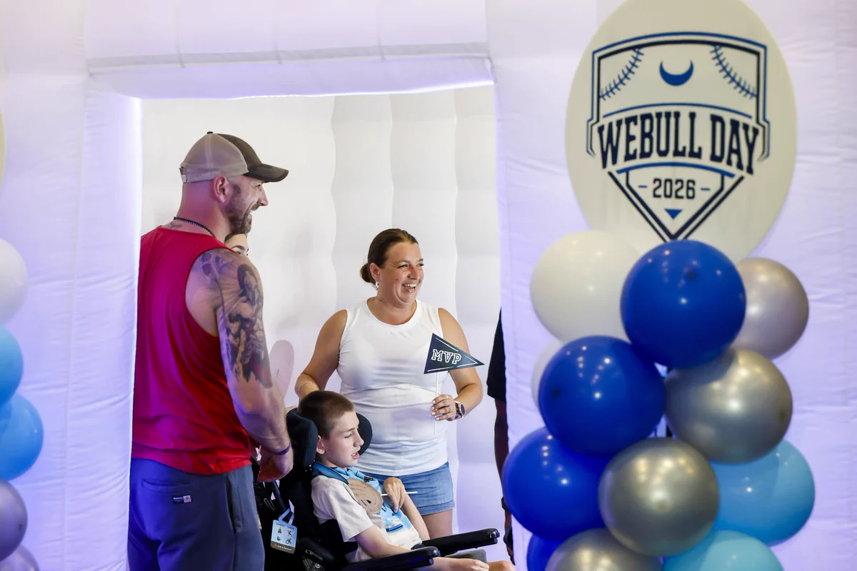 A family taking a photo in the Webull Day photo booth, next to a large Webull Day sign and blue balloon garland at Tropicana Field.