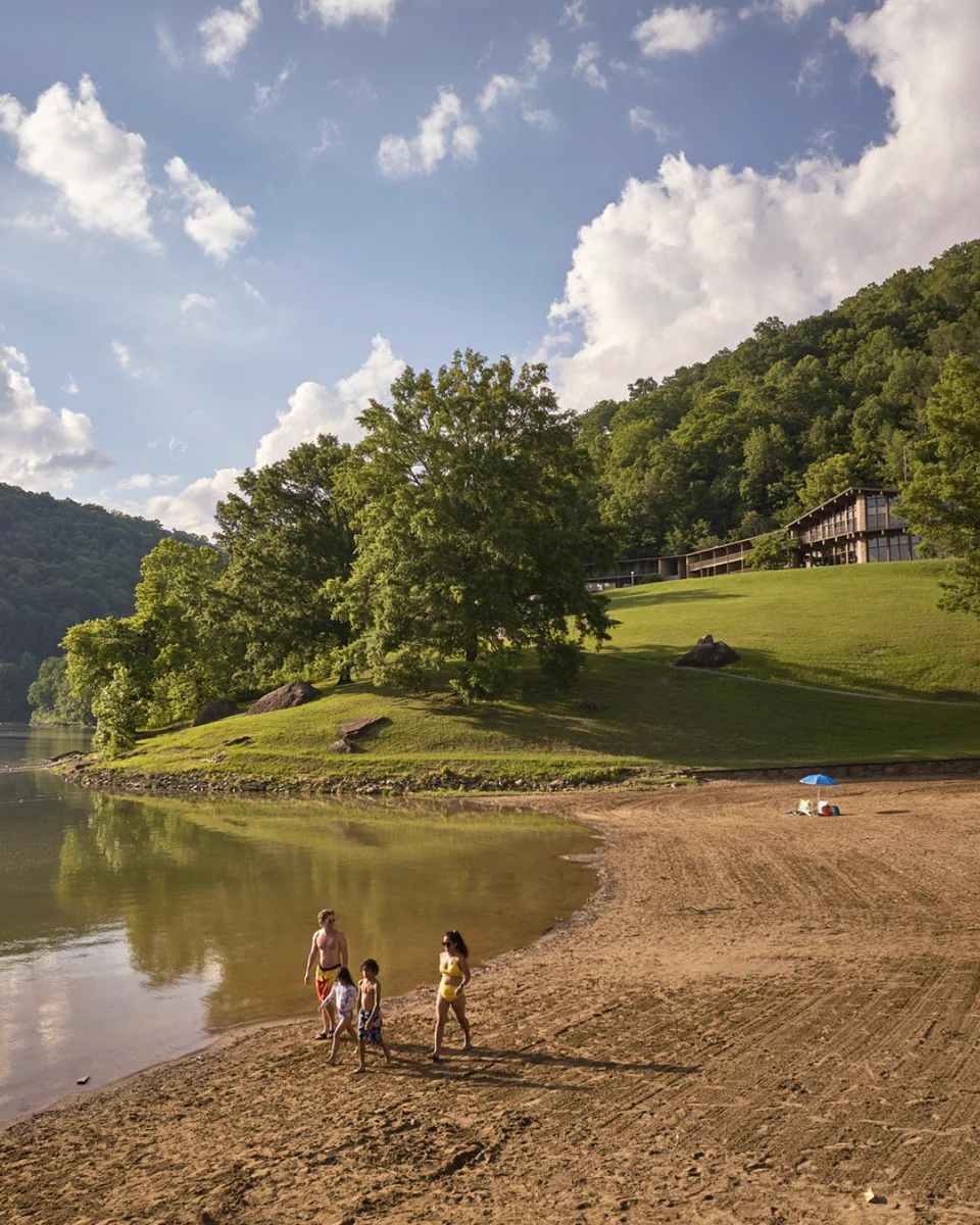 A scenic lakeside view at Babcock State Park, featuring a sandy beach with several people walking along the shore. In the background, there is a lush green hillside with a large building. The sky is partly cloudy.