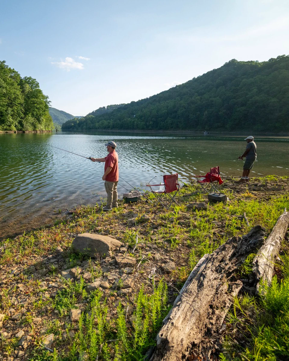 Two people fishing by the edge of a calm lake surrounded by lush green hills. They stand near some red camping chairs and fishing gear. The sky is clear and sunny.